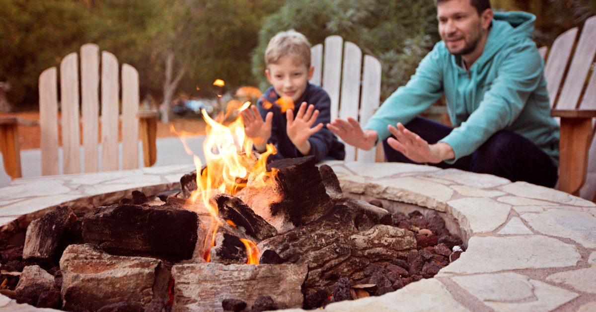 A man and a young boy sitting in wooden chairs extending their hands in front of the flame from a rock firepit.