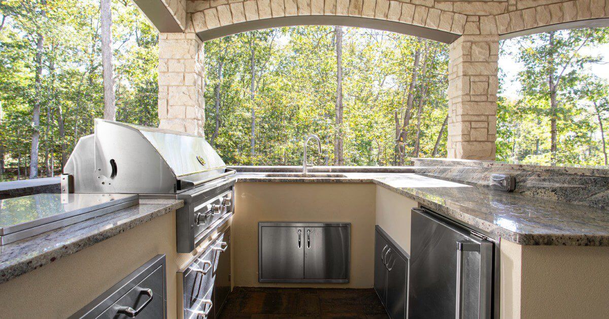 An outdoor U-shaped kitchen with stainless steel appliances, granite countertops, under a roof with rock columns.