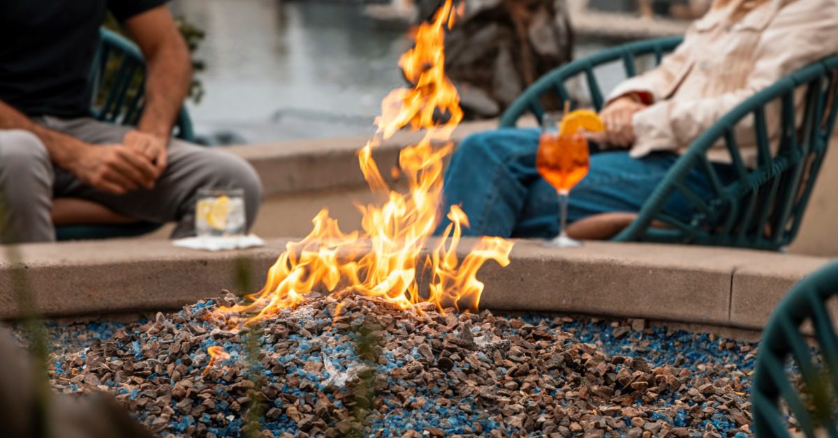 A close-up of a big flame in the center of a fire pit with a woman and a man sitting in the background with drinks.