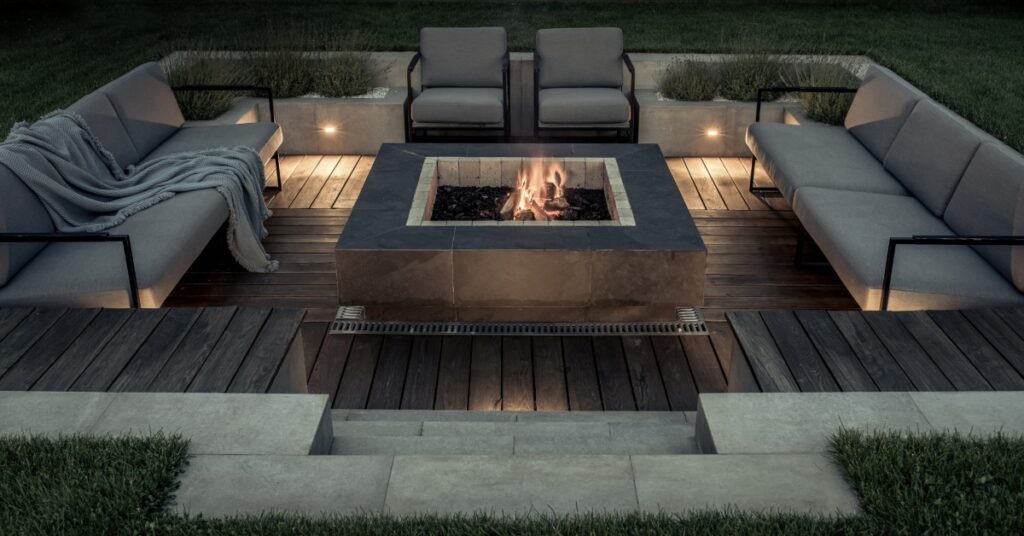 A rock square firepit in the middle of an outdoor area with couches and chairs, surrounded by grass and plants.