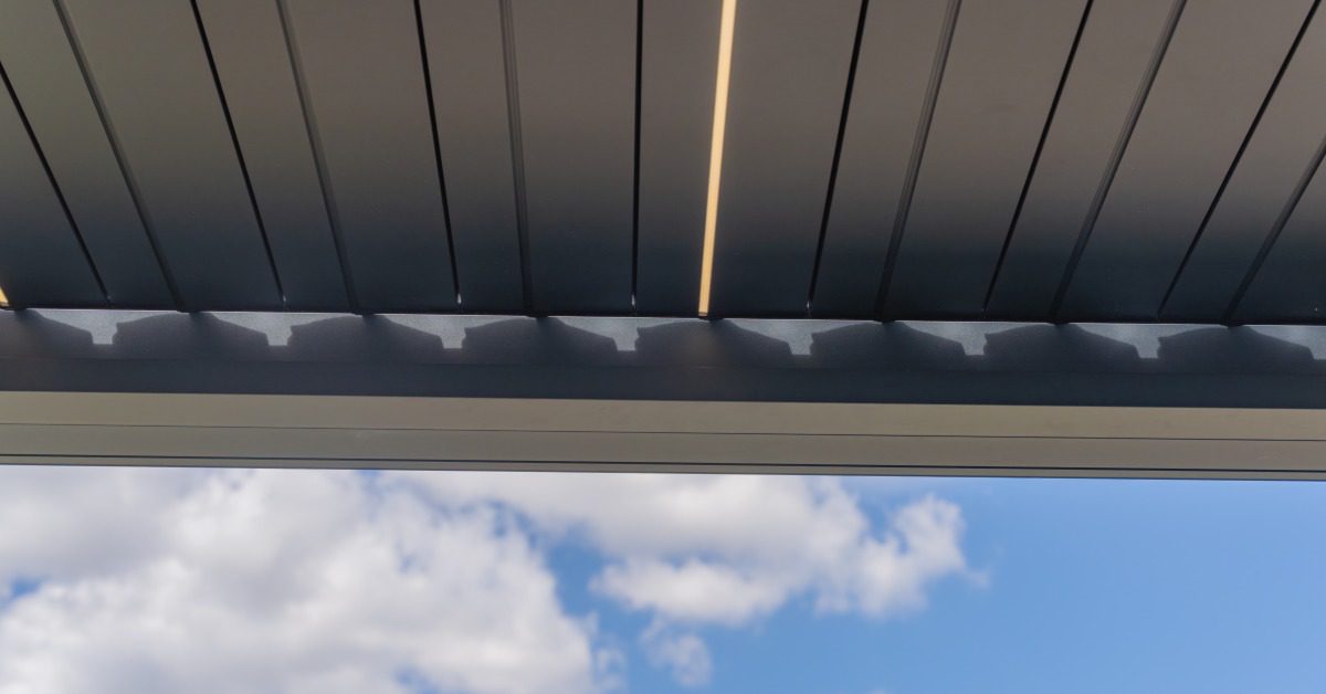 An aluminum pergola with multiple closed panels resting on a horizontal beam with multiple clouds in the background.