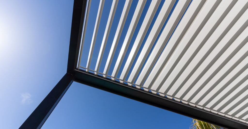 A ground view of an aluminum pergola with multiple thin white panels and thin legs, under the bright sun.