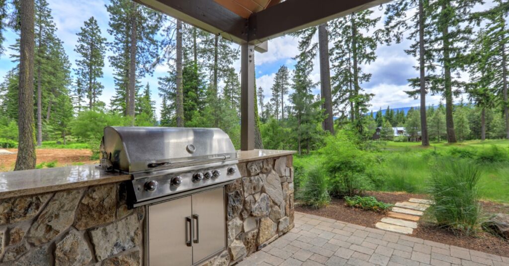 A stainless-steel grill with a large cover, built into a rock counter outdoors, under a pergola next to multiple trees.