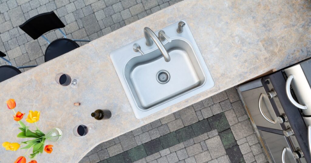 A white outdoor kitchen with black counters, a stainless-steel grill, and a sink, at the bottom of a white staircase.