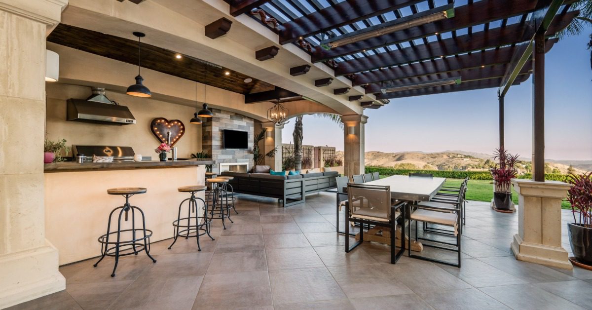 An outdoor kitchen under a large wooden pergola, with a large table, stools, and a couch with a mountain view.