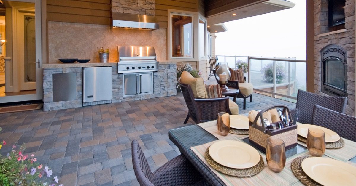 A modern outdoor kitchen with a stainless-steel grill, rock floors, a dining table, chairs, a fireplace, and a balcony.