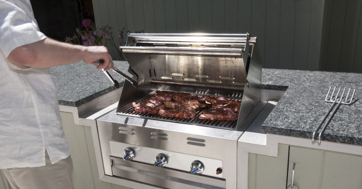 A man wearing a white shirt, standing next to a stainless-steel grill with multiple stakes, holding a pair of tongs.