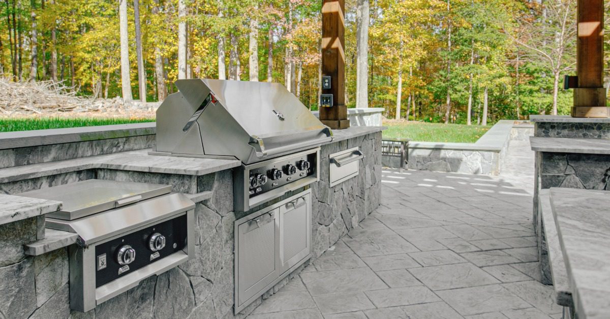 A rock outdoor kitchen with a stainless-steel grill and a cooker under a large wooden pergola next to the woods.