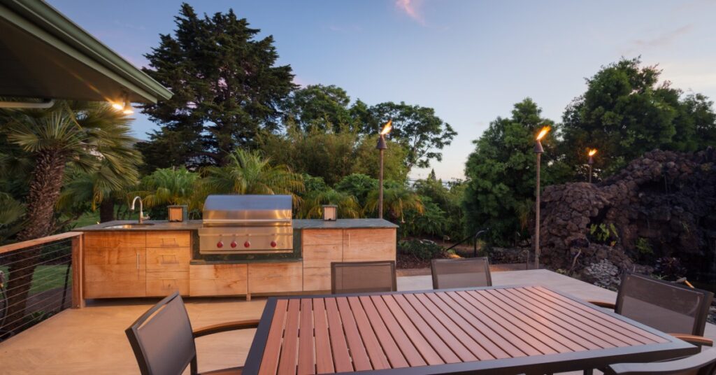 A wood dining table with six chairs outdoors next to a long counter with a stainless-steel grill and a sink.