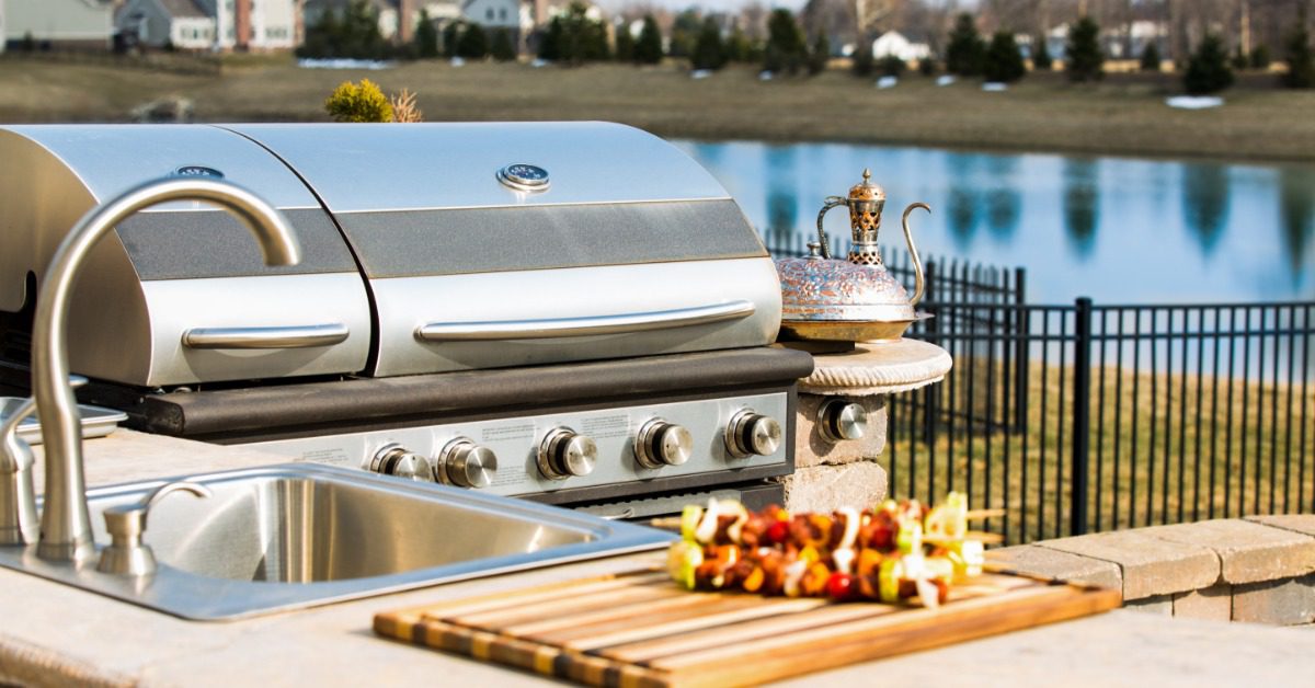 A close-up of a stainless-steel sink next to an outdoor grill placed on a brick counter with a lake in the background.