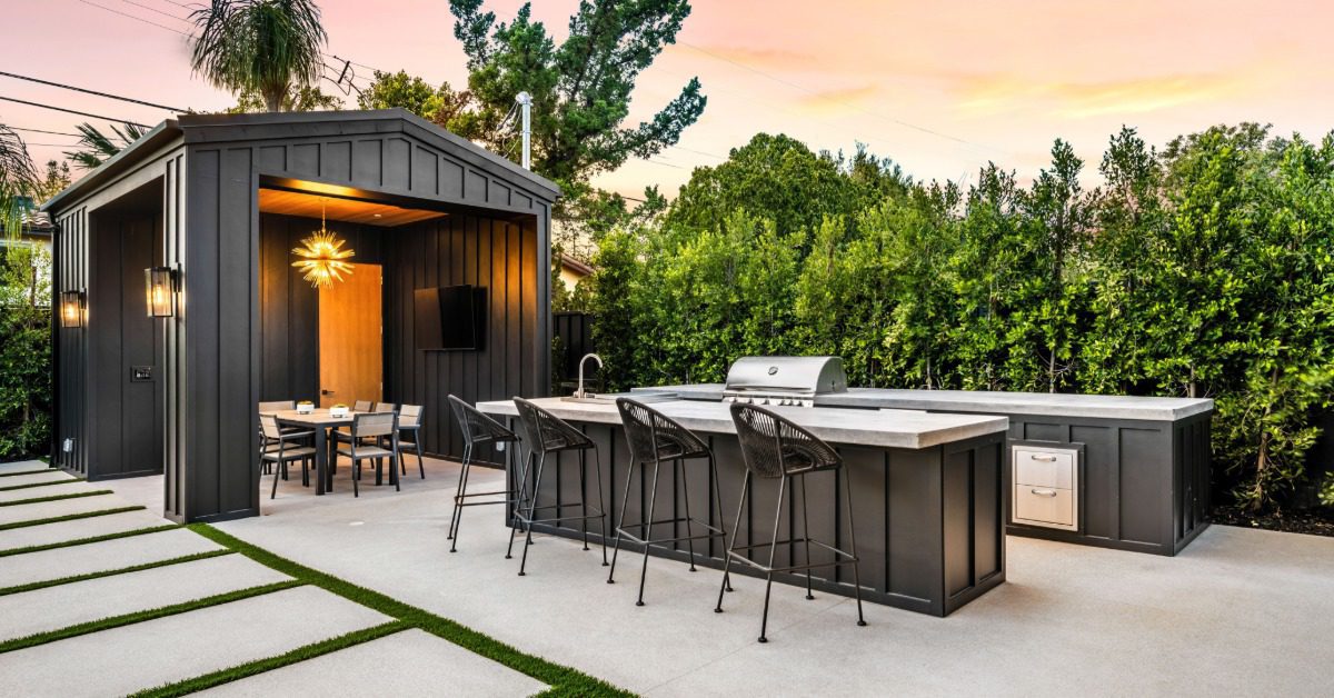 A patio with an outdoor kitchen, a counter with four chairs and a dining table inside a small shed with a chandelier.