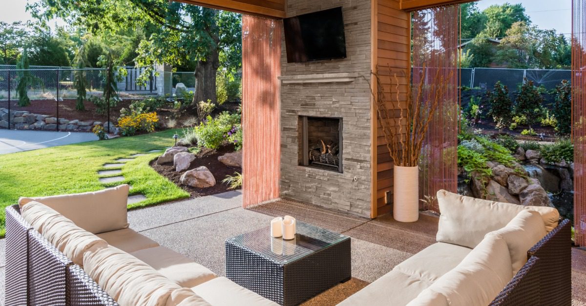 A wooden pergola with a rock column holding a flat tv screen and a fireplace, over a sectional couch on a yard.