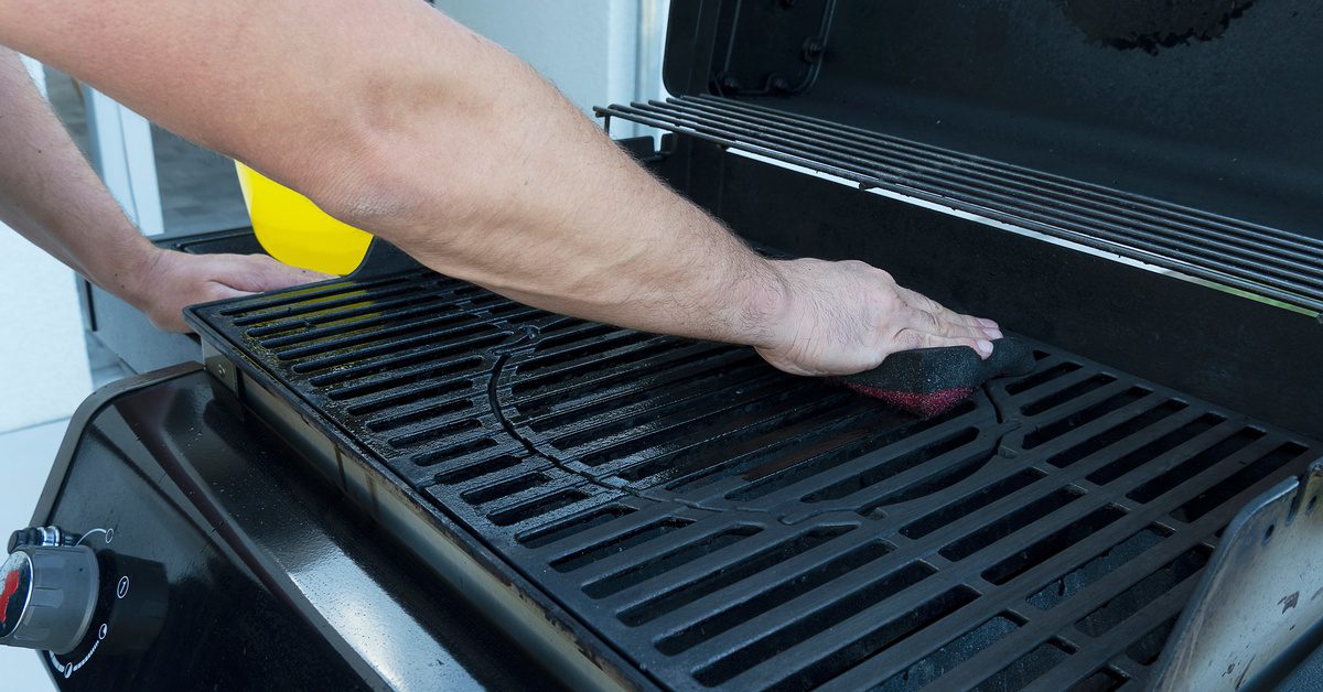 A close-up of an arm using a sponge to clean the interior of a metal grill with stainless-steel knobs and a cover.