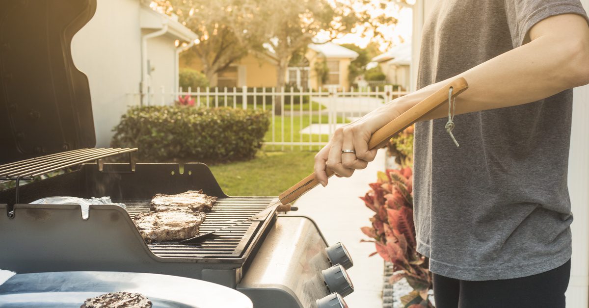 A man holding a long wooden handle spatula on a hamburger placed on a grill outdoors next to a plate full of meat.