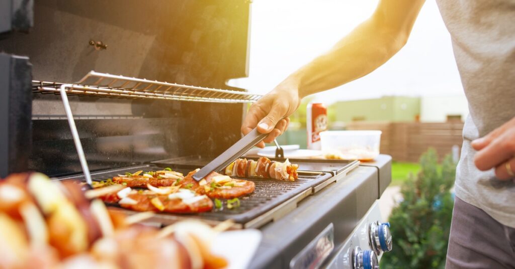A close-up of the hands of a man using a pair of tongs to grab a piece of meat on a grill outdoors during a sunny day.