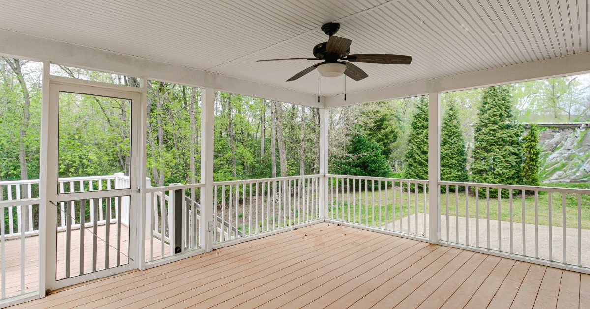 An enclosed space with white columns and a white ceiling, a brown ceiling fan, and a safety rail surrounded by trees.