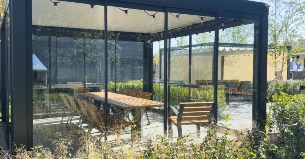 A metal pergola covered in glass panels with string lights hanging inside and a long wooden table with eight chairs.