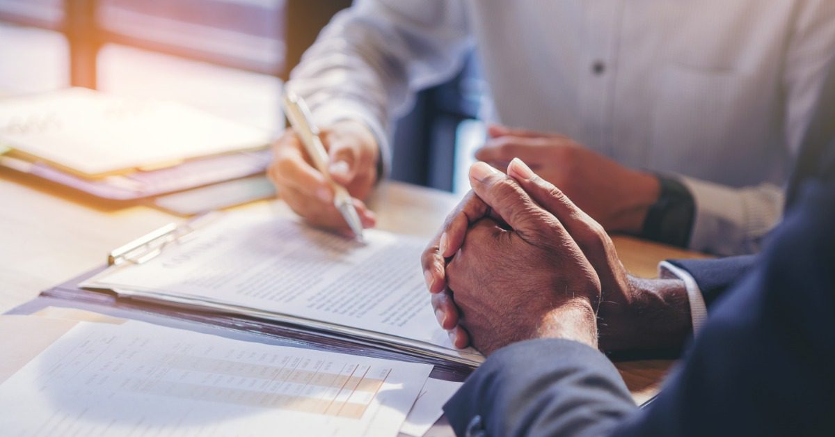 A close-up of a hand holding a pen and signing a document on a desk, next to a man waiting with crossed hands.