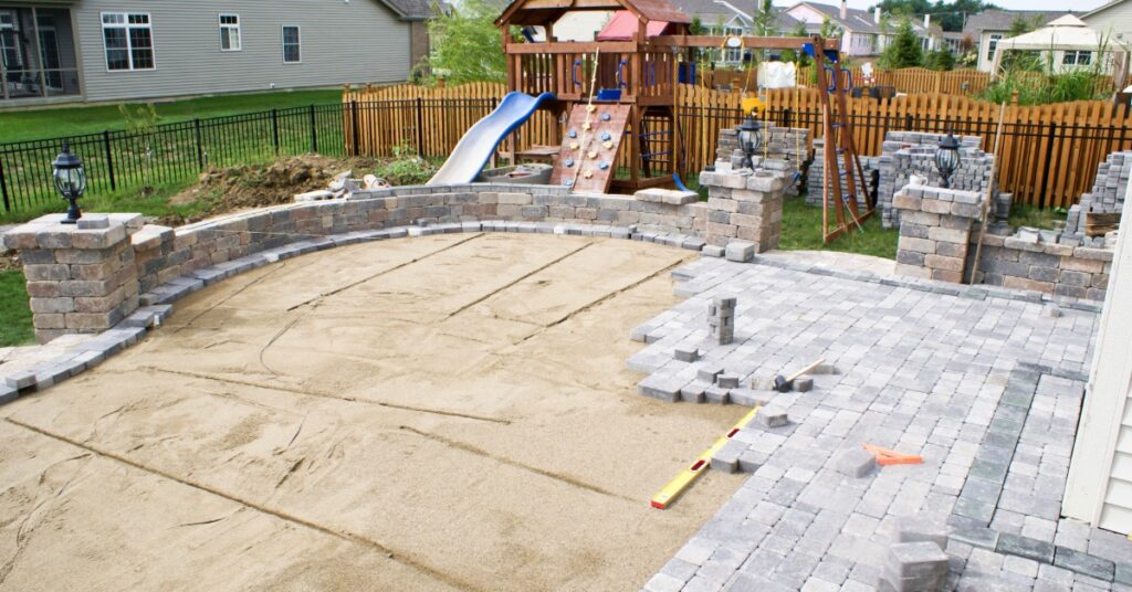 A large patio with an unfinished floor and multiple concrete blocks piled on the side next to a children’s playground.