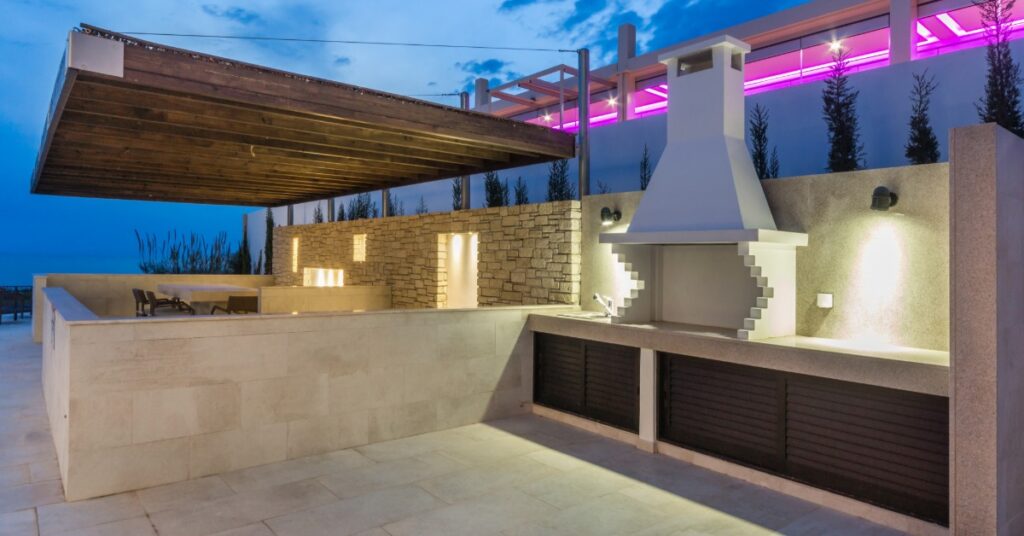 An outdoor kitchen with marble floors, a stove with a large chimney, and a counter next to a large sitting area.