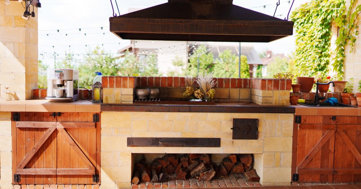An outdoor kitchen with a large stone oven, a sturdy chimney, wooden storage doors, and neatly stacked firewood below.