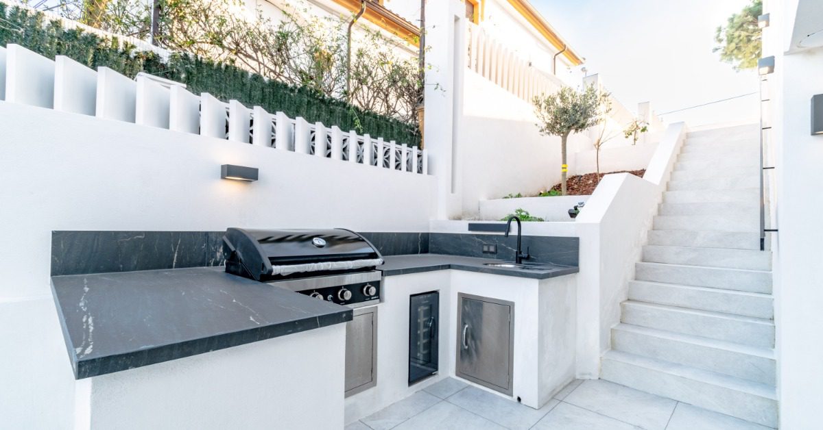 An outdoor kitchen with granite countertops, a stainless-steel grill, and a faucet, next to a white concrete staircase.