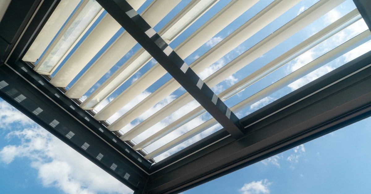 A ground view of an aluminum pergola with multiple angled white panels on the top blocking the sun partially.