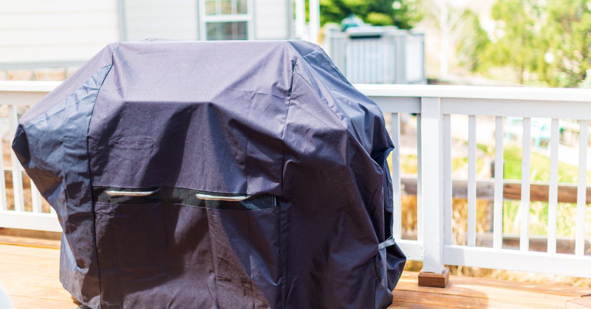 An individual grill placed on a wooden deck with a white rail, covered with a black cloth under bright sunlight.