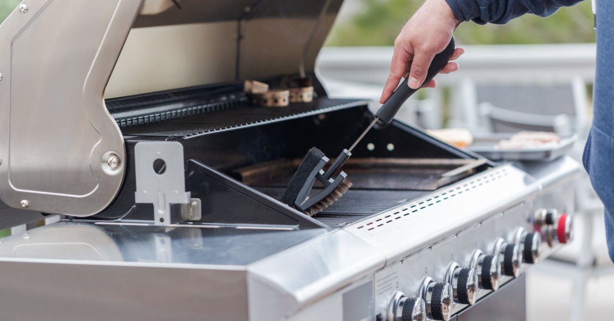 A close-up of a hand holding a brush to clean the grates of an outdoor stainless steel grill with multiple knobs.