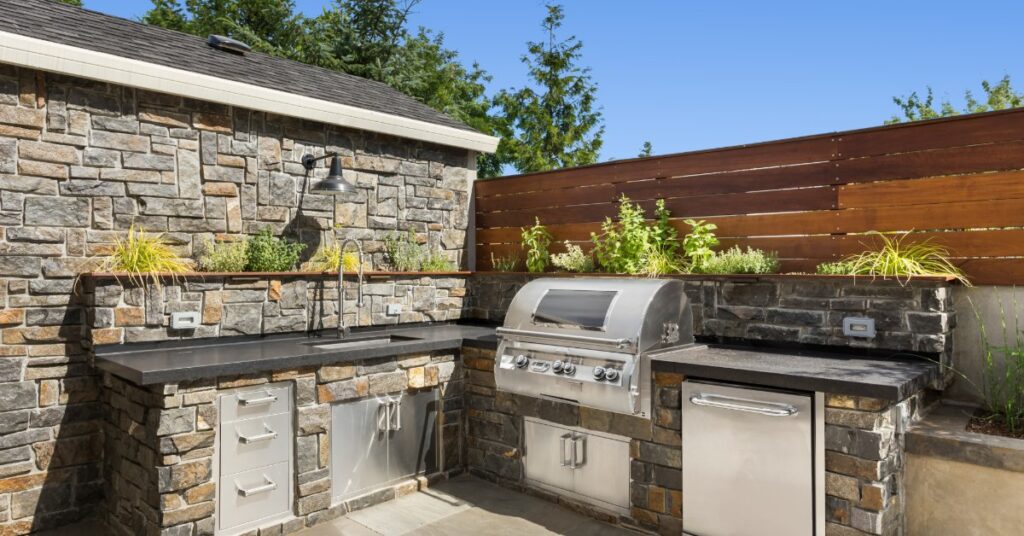 A rock outdoor L-shaped kitchen with a stainless-steel grill, a small refrigerator, and a sink on a sunny day.