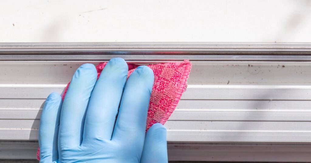 A close-up of a hand wearing a blue latex glove, using a pink sponge to clean the side of a white aluminum cover.