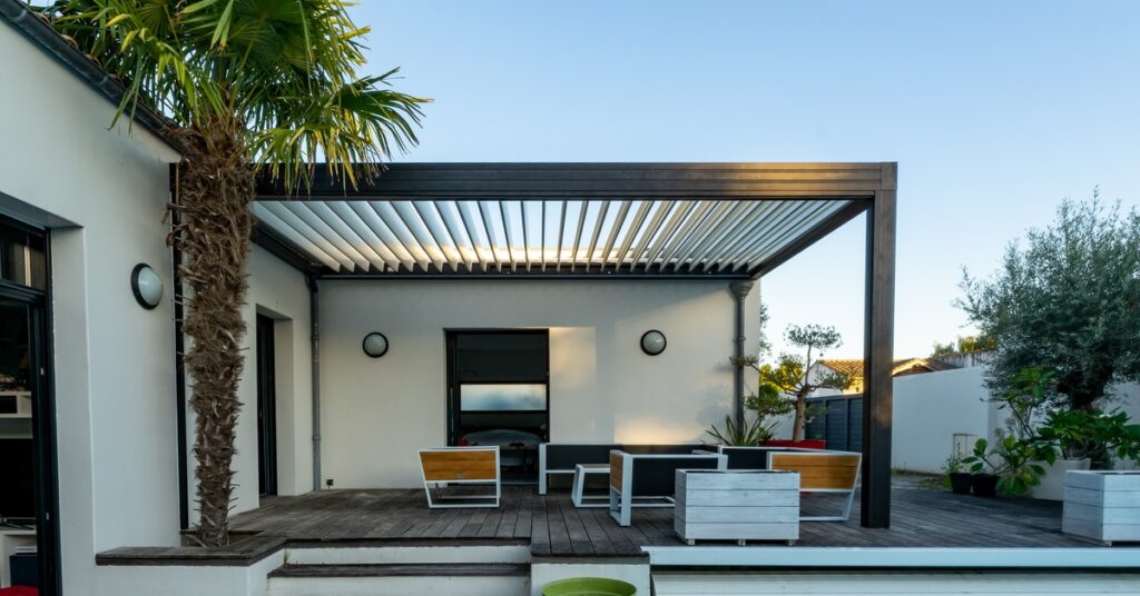 An aluminum cover with plastic panels over an outdoor deck with a sitting area, a palm tree and multiple plants.