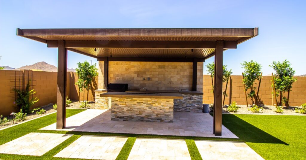A wooden patio cover over an outdoor kitchen with an island and stainless steel appliances surrounded by grass.