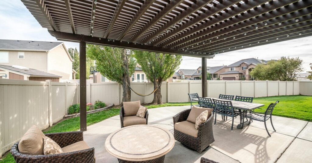 A wooden patio cover with multiple panels over a sitting area with four woven chairs and a metal dining table.