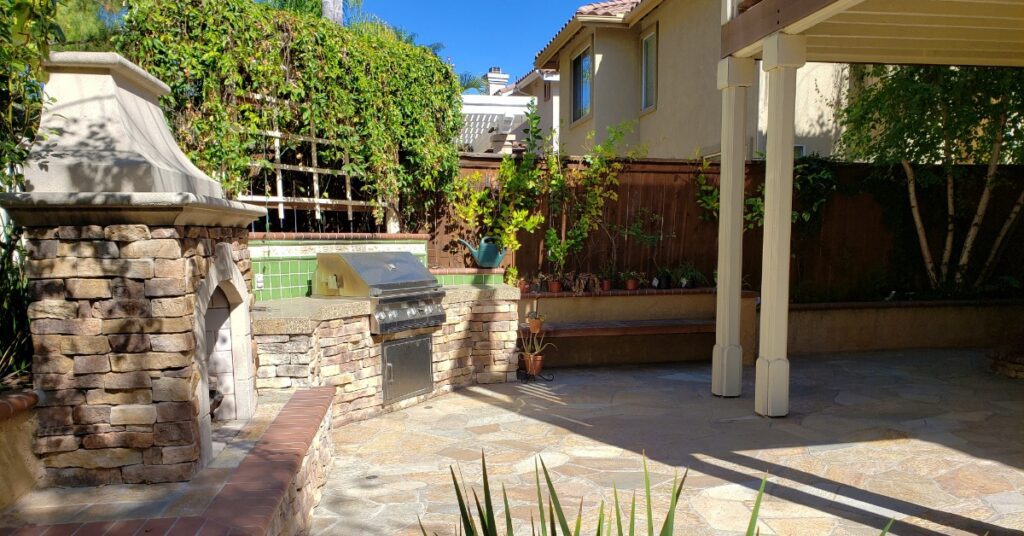 A backyard with tiled floors, a pergola, and a stainless steel built-in grill next to a wood oven and a bench.