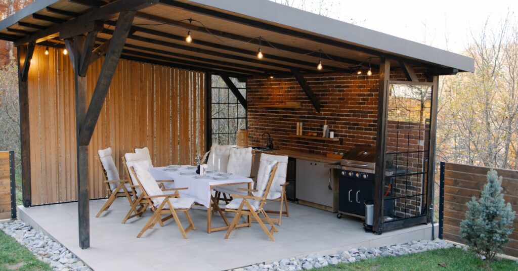 An outdoor kitchen next to a large dining table, under a pergola with hanging string lights over a concrete platform.