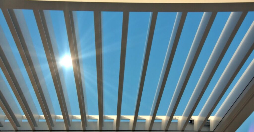 The top of a white aluminum pergola with multiple vertical moving panels letting the sun shine through them.