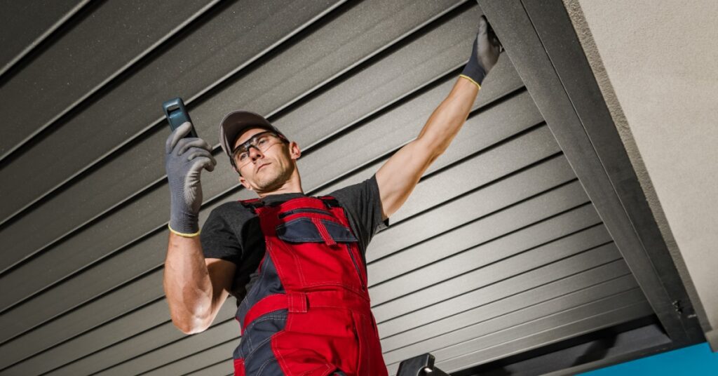 A worker wearing red overalls and safety glasses, standing on top of a ladder touching the roof of an aluminum pergola.