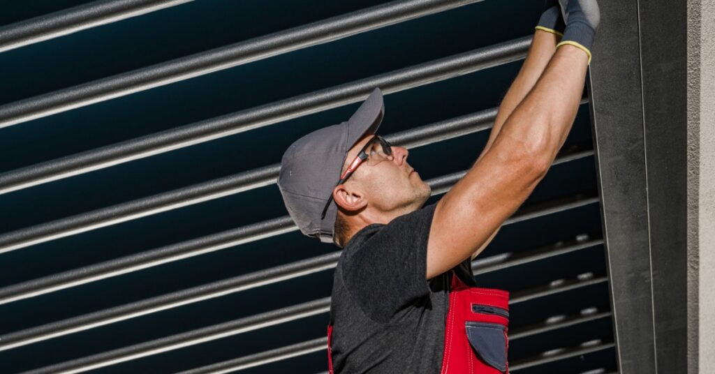 A worker wearing black gloves touching the interior edge of an aluminum pergola with multiple tubes on the ceiling.