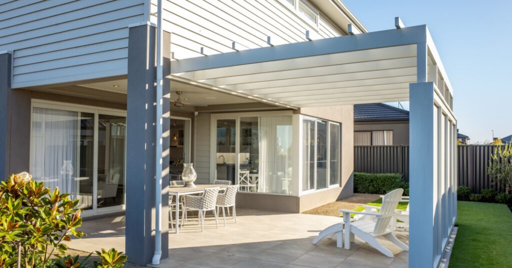 A two-story home with a large outdoor space under a white pergola with two lounging chairs and a dining table.