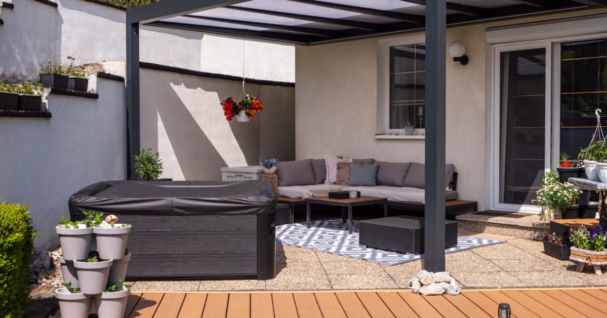 A garden with a wooden walkway, potted plants, a covered hot tub, and a sectional couch under an aluminum pergola.