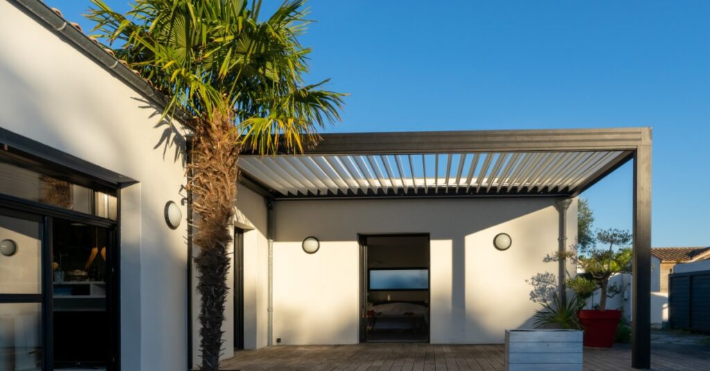 A white one-story home with an aluminum pergola on the side next to a large palm tree over a wooden deck.
