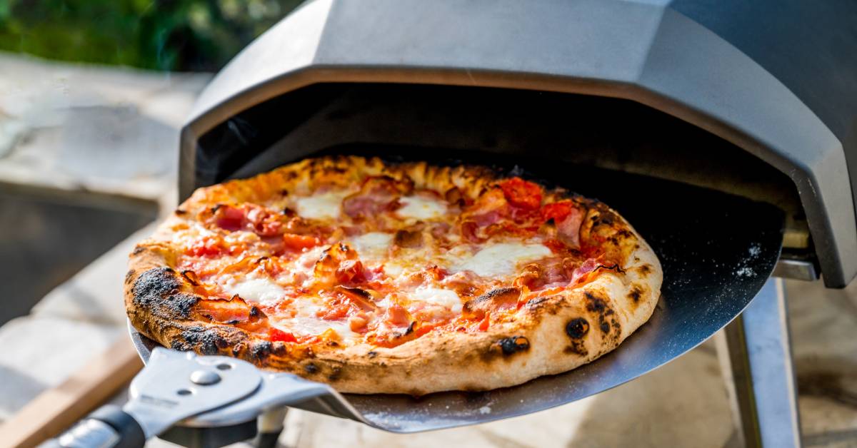 A close-up of a pepperoni pizza with burnt edges on a large metal tray next to a small freestanding oven.