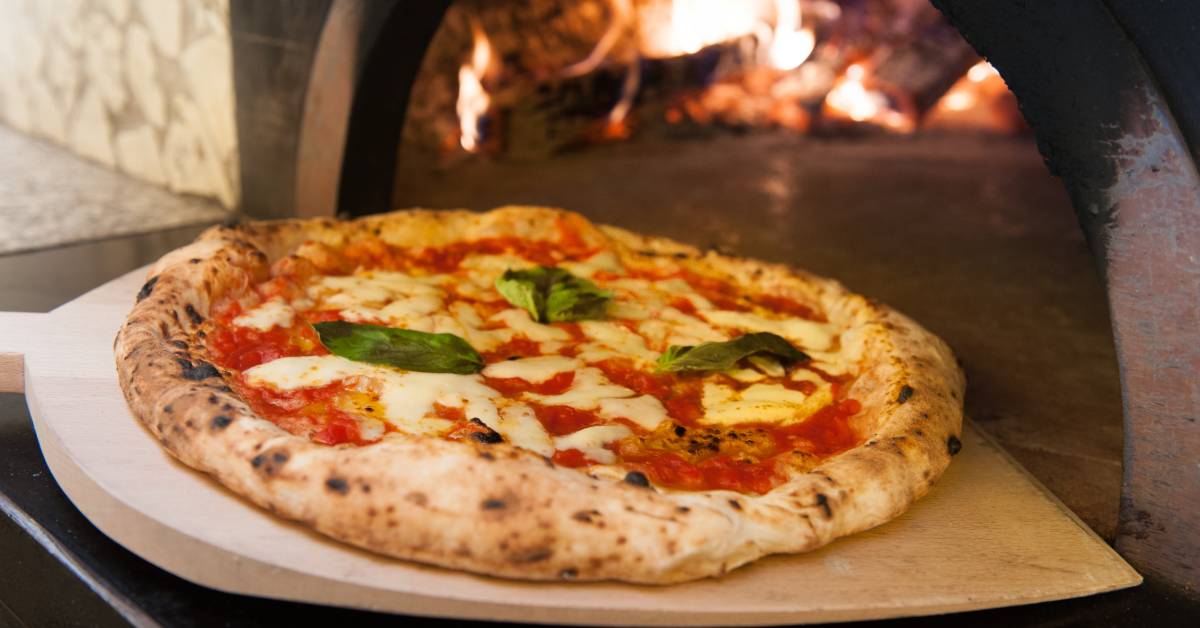 A close-up of a Margherita pizza on a wooden tray placed next to a large rock oven with burning wood inside.