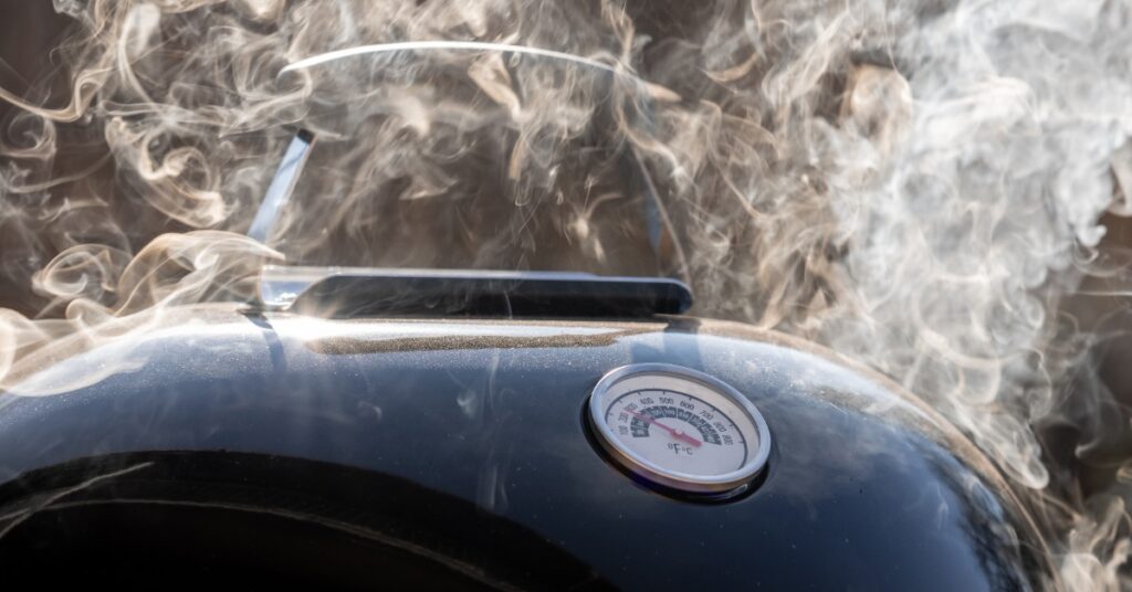 Close-up of a grill with a black top and a built-in thermometer with a large cloud of smoke coming from inside.