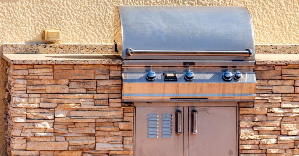 A stainless-steel grill with a rounded cover, knobs, and a storage compartment underneath, placed on a rock counter.