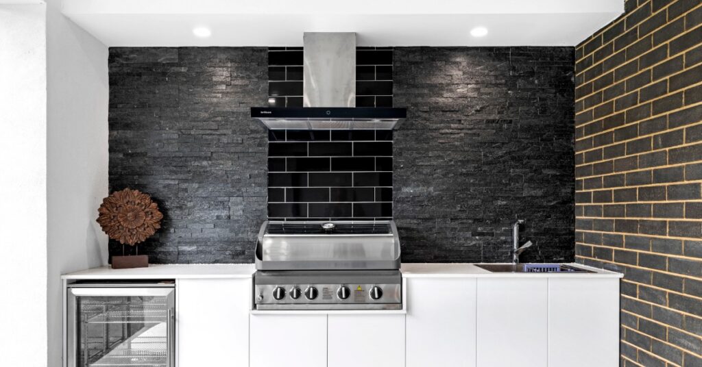 A white outdoor kitchen with a black backsplash wall and a stainless-steel grill between a small fridge and a sink.