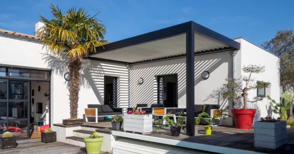 A one-story home with an outdoor deck covered with an aluminum pergola over outdoor couches, next to a palm tree.
