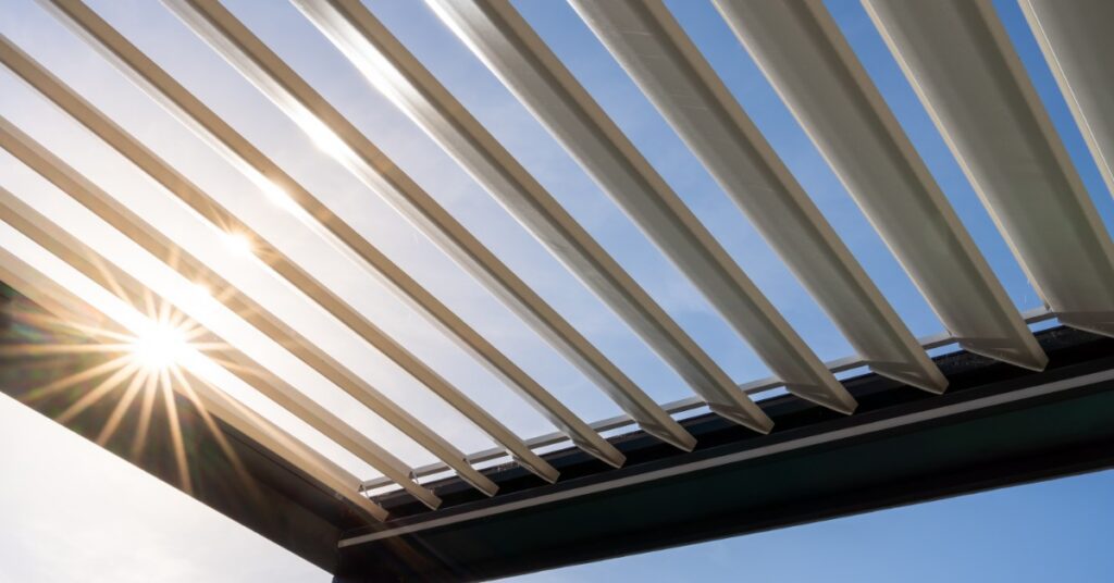 A close-up of the top of an aluminum pergola with white open moving panels, with the sun shining brightly through them.