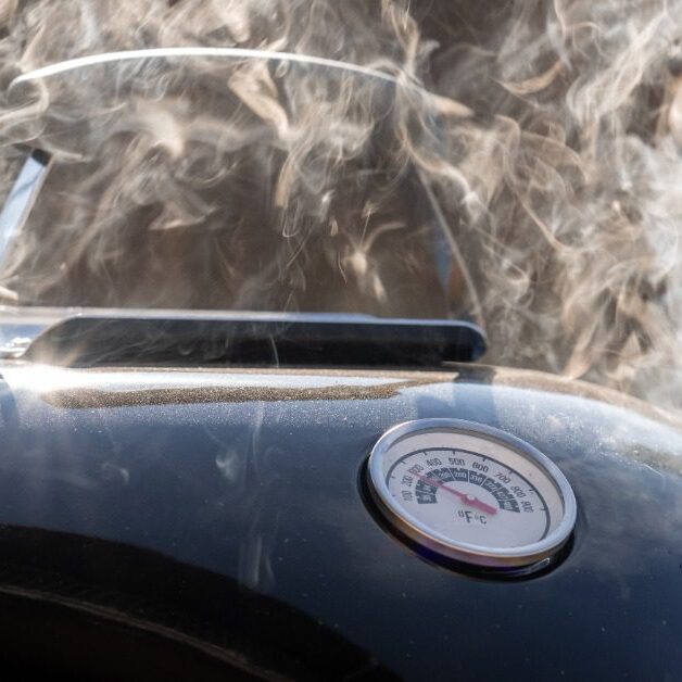 Close-up of a grill with a black top and a built-in thermometer with a large cloud of smoke coming from inside.